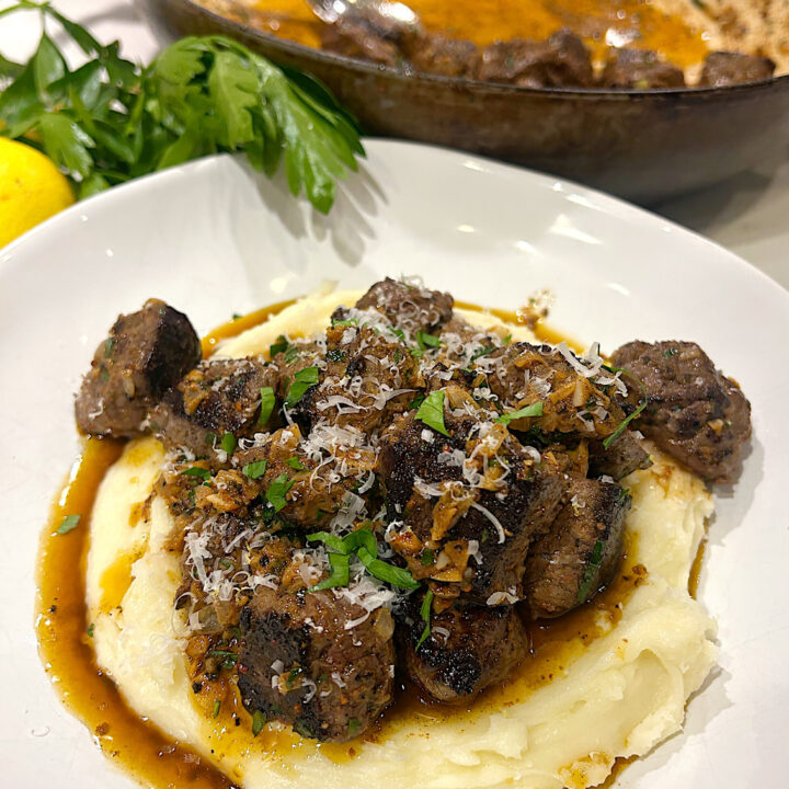 a white bowl of mashed potatoes topped with steak bites and a buttery sauce
