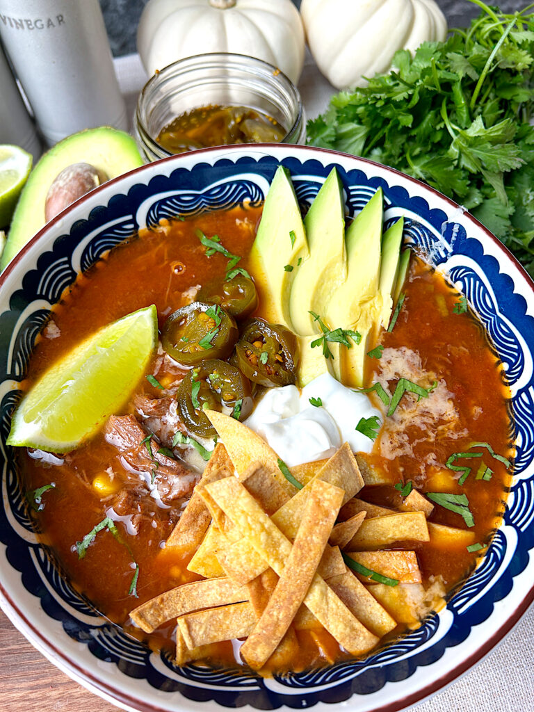 a blue and white bowl of tomato based soup full of corn, black beans and meat, topped with avocado, lime and tortilla strips