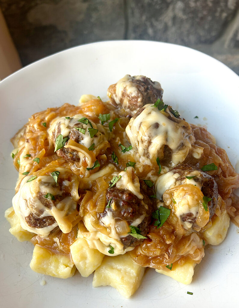 a white bowl of gnocchi topped with caramelized onions, meatballs and Swiss cheese
