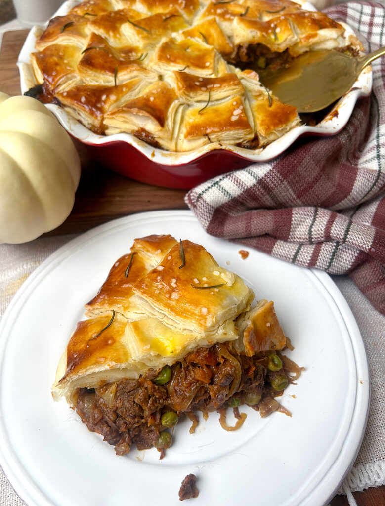 a white plate with a slice of meat pie with flaky pastry on top
