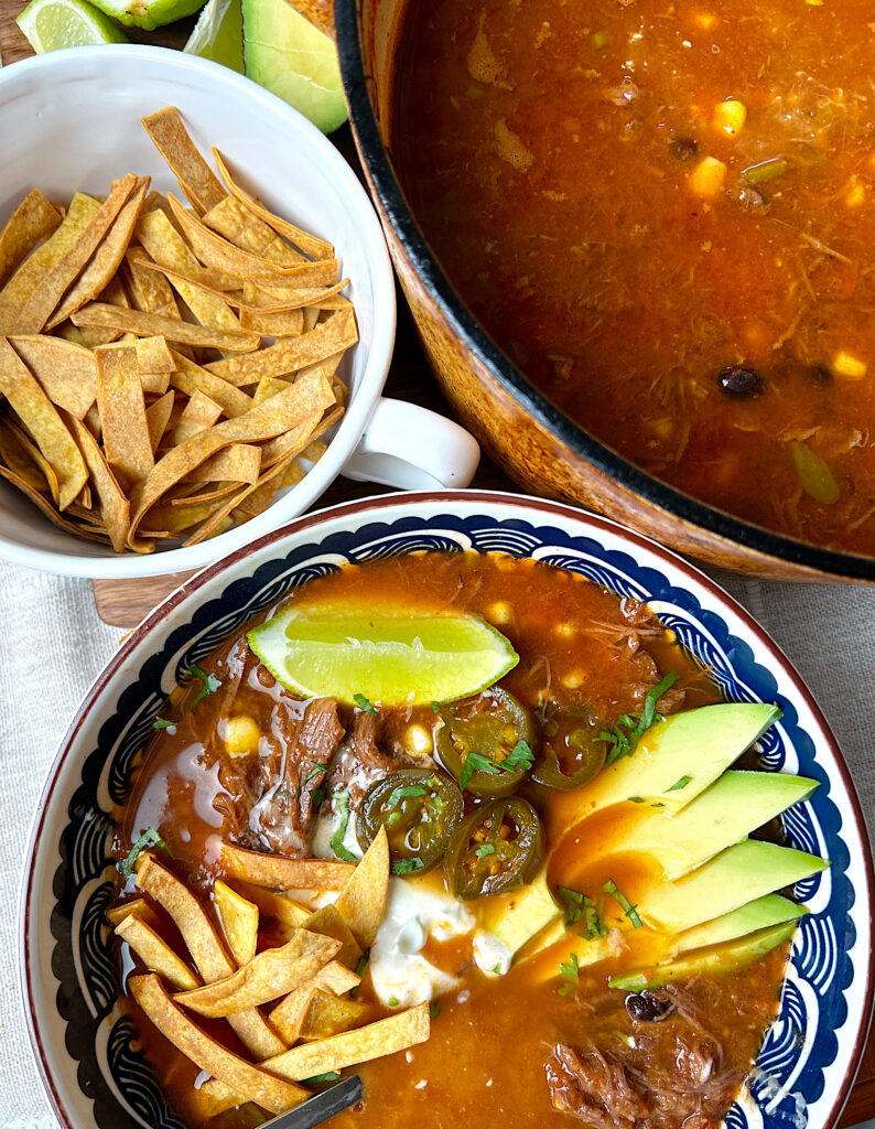 a pot of tortilla soup with a blue bowl full of soup next to it topped with avocado, tortillas and sour cream