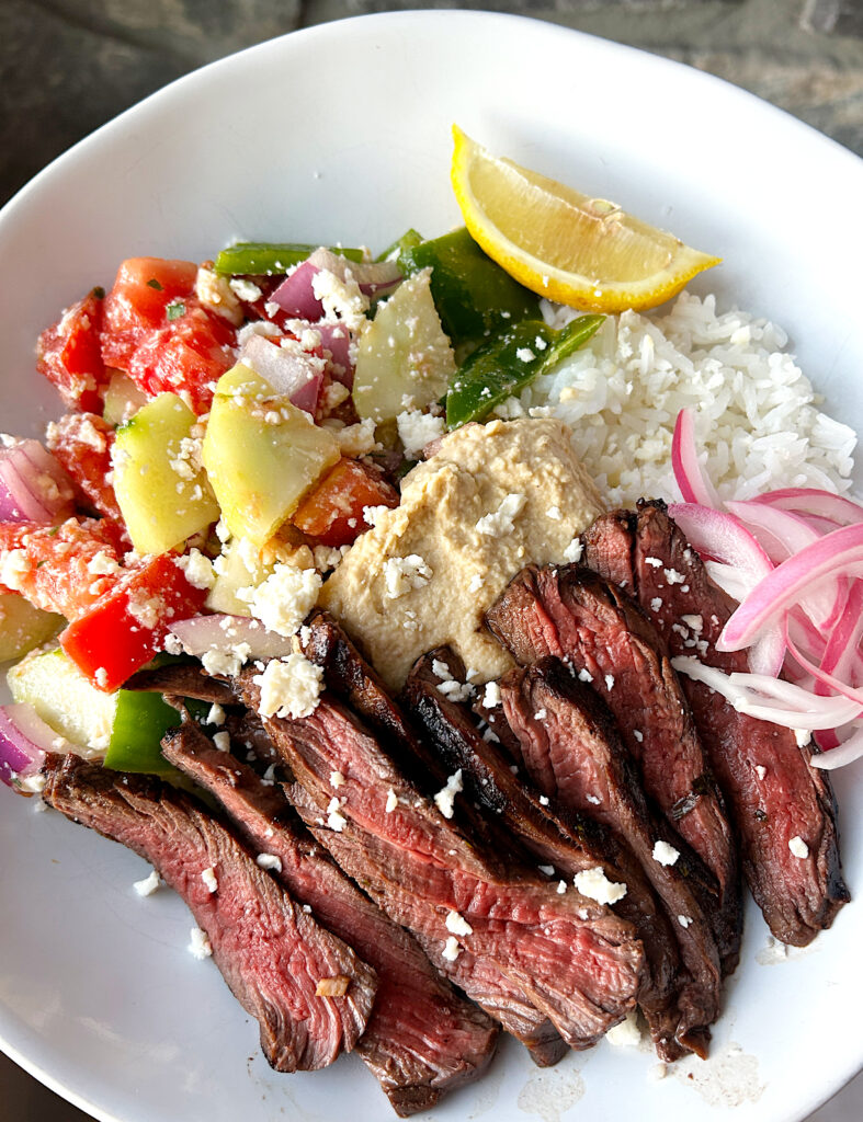 a white bowl with rice, greek salad, sliced steak and pickled onions