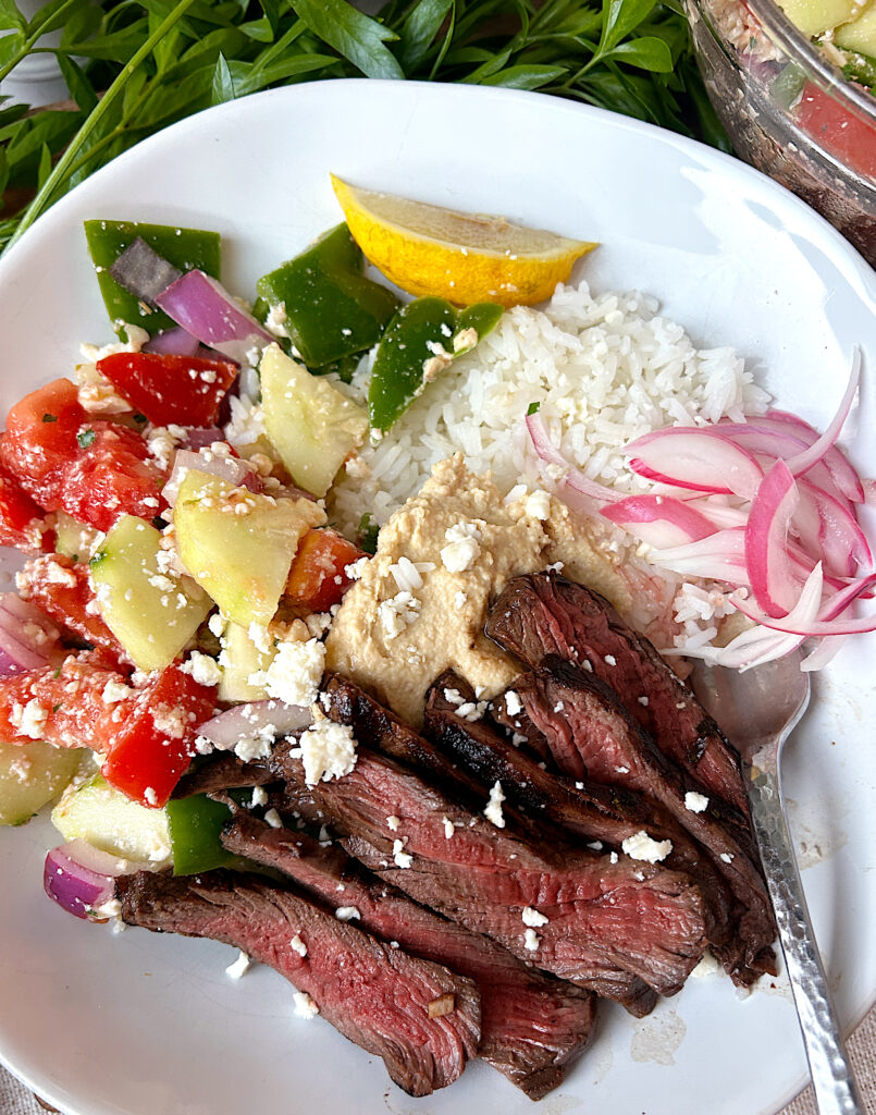 a white bowl of rice, greek salad, venison steak and hummus